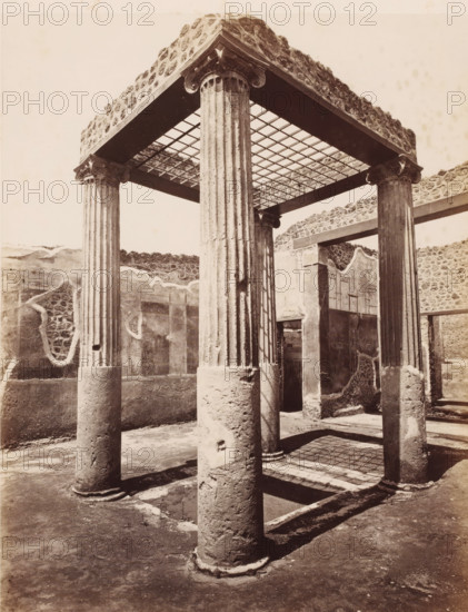 Remains of a shop in Pompeii, Italy, photograph, number: '10', number: '34.', inscription: 'Pompei interior d'une boutique fouilles 1877', Pompeii, 1877 - 1900, photographer, pasteboard, paper, albumen print, albumendruk, height 307 mm x width 405 mm, ruin of a building, architecture, A sepia-toned photograph of an ancient architectural ruin showing a square canopy supported by fluted stone columns with carved capitals, a grid-like ceiling above casting a shadow pattern on the ground, surrounding low walls with patches of rough stonework and plaster, an open doorway set into a wall, and a worn earthen floor with column bases and long shadows stretching across it.