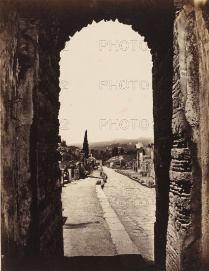 View through the gate of Herculaneum in Pompeii, Italy, photograph, inscription: 'Pompei porte Herculaneum, squelette faction', number: '20.', number: '10', Pompeii, 1851 - 1900, photographer, pasteboard, paper, albumen print, height 404 mm x width 305 mm, ruin of a building, architecture, gate, entrance, A view through a large, rough stone archway onto a narrow paved street flanked by ancient stone walls and ruins, with a cobbled path running down the center and a raised sidewalk on one side, scattered stones and fragments along the edges, a tall slender cypress tree visible in the distance against a pale sky, distant low hills on the horizon, and weathered building facades and column remnants lining the street.