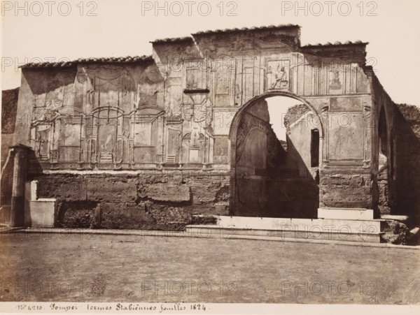 Termes Stabiennes fouilles 1824, Ruin of the Stabian Baths in Pompeii, Italy, Pompei, photograph, number: '21', number: 'No. 4215', number: '57', number: '11', photographer: attributed to Giorgio Sommer, Pompeii, 1857 - 1875, Attributed to, pasteboard, paper, albumen print, albumendruk, height 307 mm x width 405 mm, ruin of a building, architecture, excavation, archeology, A wide monochrome photograph of a weathered stone wall with architectural fresco panels and molded decorations, featuring a large rounded archway at one side revealing a narrow passage beyond and casting a diagonal shadow, a rectangular niche above the arch containing a sculpted relief of a seated figure, carved frames and drapery motifs across the wall surface, a tiled roofline with uneven eaves, rough exposed masonry at the lower portion of the wall, a stepped white stone base in front, and a flat dirt foreground extending to the bottom edge of the image.