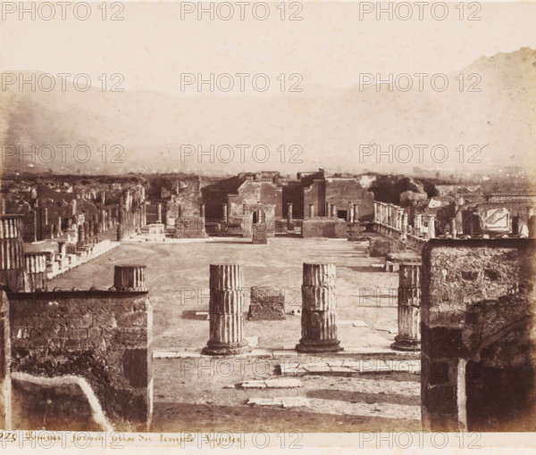 Forum prize du temple Jupiter, View of the Forum from the Temple of Jupiter in Pompeii, Italy, Pompei, photograph, number: '10', number: '16.', number: '266', photographer: attributed to Giorgio Sommer, Pompeii, 1857 - 1875, Attributed to, pasteboard, paper, albumen print, height 308 mm x width 405 mm, square, circus, ruin of a building, architecture, Temple of Jupiter (Pompeii), A sepia-toned aerial view of an ancient ruin site showing weathered stone columns, some freestanding and some in short stacked sections, arranged on a paved ground with rectangular stone slabs and low masonry platforms; flanking walls and partial structures with arched openings and square doorways stand toward the background, interspersed with rows of smaller columns and ruined facades; the foreground shows thick textured stone walls framing the scene and truncated column drums positioned near the viewer; beyond the ruins a hazy landscape of low hills and distant mountains rises under a pale sky.
