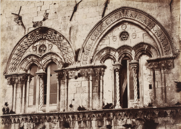 Gothic windows of the Palazzo Montalto in Syracuse, Sicily, Finestre gothique del Palazzo Montalto (Siracusa), photograph, number: 'No. 1377', number: '22', photographer: Giorgio Sommer, Syracuse (city), 1857 - 1914, pasteboard, paper, albumen print, height 312 mm x width 386 mm, open window, parts of church exterior and annexes, A close view of a weathered stone facade showing two adjacent arched window openings with ornate carved surrounds and decorative columns; the left arch features a zigzag chevron pattern and a small circular floral motif above paired narrow windows with twisted and smooth shafts, while the right arch displays a leaf and geometric border with a round tracery opening above three narrow arches and a pair of twisted columns flanking a recessed doorway; the stone surface shows darkening and mossy patches, chipped edges, and layered masonry blocks with a horizontal carved cornice and a row of triangular leaf-like decorations along the lower band.