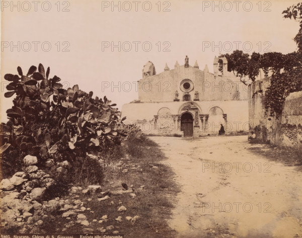 Entrance to the catacombs of the Chiesa di San Giovanni in Syracuse, Italy, Chiesa di S. Giovanni. Entrata alle Catacombe, Siracusa, photograph, number: '1805.', Syracuse (city), 1851 - 1900, photographer, Sicily, pasteboard, paper, albumen print, height 200 mm x width 251 mm, church (exterior), catacombs, A sepia-toned photograph showing a worn stone building with an arched doorway and decorative round window above, flanked by small statues and architectural details, set behind a dirt path leading toward it; to the left foreground there is a dense patch of broad flat-leaved plants and scattered rocks, while to the right a low wall and overhanging trees frame the scene and a solitary figure in dark clothing sits near the building entrance.