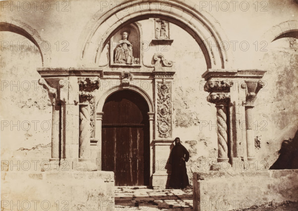 Entrata delle Catacombe in Siracusa, Entrance to the catacombs in Syracuse, Sicily, photograph, number: 'No. 1386.', photographer: Giorgio Sommer, Syracuse (city), 1857 - 1914, pasteboard, paper, albumen print, height 309 mm x width 385 mm, gate, entrance, catacombs, A stone building facade with a large arched doorway framed by carved columns and decorative reliefs, a recessed niche above the doorway containing a seated sculpted figure, spiral-fluted columns and ornate capitals on either side, weathered plaster and stone surfaces with visible cracks and patches, a tall dark wooden door within the arch, stone steps and a low wall in front, and a person dressed in a dark full-length garment standing near the doorway.