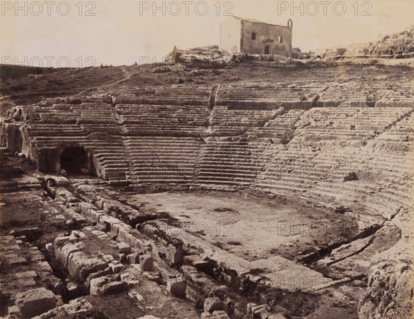 View of the ruins of the Greek amphitheater in Syracuse, Sicily, Siracusa Teatro greco, photograph, number: '2667', photographer: Giorgio Sommer, Syracuse (city), 1857 - 1914, paper, albumen print, albumendruk, height 198 mm x width 249 mm, ruin of a building, architecture, amphitheater, A wide view of an ancient stone amphitheater with concentric carved seating tiers leading down to a flat central area, the worn stone showing grooves and cracks, stair-like divisions and a dark arched opening set into the lower seating; a rough rocky foreground with scattered blocks and channels runs toward the stage, and a grassy terrace with uneven paths rises behind the upper seats where a simple rectangular stone building with small window openings and a doorway sits atop the hill.
