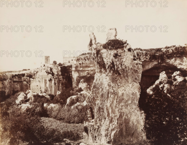 Latomie del Paradiso in Siracusa, View of Latomie del Paradiso in Syracuse, Sicily, photograph, number: 'No. 1384.', photographer: Giorgio Sommer, Syracuse (city), 1857 - 1914, pasteboard, paper, albumen print, height 309 mm x width 385 mm, quarry, A sepia-toned landscape showing a rugged cliff face with rough, weathered rock formations rising prominently in the center and right, textured with crevices and patches of vegetation clinging to ledges. In the midground a tall, narrow pillar of rock stands apart from the main cliff, topped with low shrubs, and a flat-topped ruin sits further back on the plateau. Dark cave-like openings and overhangs are visible along the cliff line, while scattered boulders and dense low bushes occupy the foreground, creating a contrast between the pale limestone surfaces and the darker foliage. The sky above is pale and largely empty.