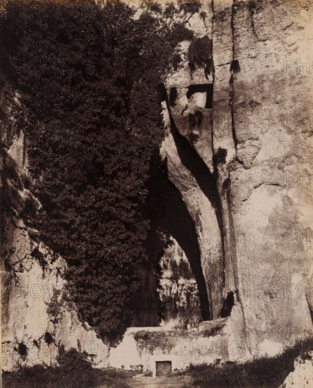 View of the Ear of Dionysius in Syracuse, Sicily, Orecchio di Dionisio, Siracusa, photograph, number: '2664', photographer: Giorgio Sommer, Syracuse (city), 1857 - 1914, pasteboard, paper, albumen print, height 254 mm x width 202 mm, cave, grotto, Ear of Dionysius, A tall vertical rock face fills the scene with rough textured stone on one side and a dark shadowed crevice on the other, a large overhanging ledge creating deep contrast where the rock curves inward; dense foliage clings to one portion of the cliff, forming a dark mass of leaves and branches against the lighter stone; beneath the overhang a small rectangular doorway set into the rock is visible with a flat area of ground leading up to it and low vegetation along the base of the cliff.