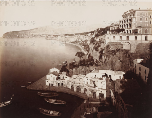 Sorrento, View of Sorrento, Italy, photograph, number: '6189', photographer: Giorgio Sommer, 1857 - 1914, pasteboard, paper, albumen print, albumendruk, height 308 mm x width 386 mm, city-view in general, 'veduta', gulf, bay, A sepia-toned coastal panorama showing a calm bay with small boats moored near a curved shoreline, whitewashed buildings with arched openings and tiled roofs clustered on a rocky promontory, steep cliffs rising behind the buildings topped with trees and terraces, a multi-story villa with balconies and arched colonnades perched at the cliff edge, a stone pier and wooden platforms extending into the water, and distant low mountains along the horizon under a pale sky.