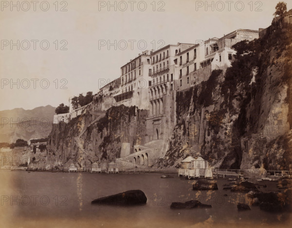 View of the cliffs of Sorrento and Hotel Tramontano, Italy, Hôtel Tramontano, Sorrento, photograph, inscription: 'Sorrento Hotel Tramontano', number: '2127', photographer: Giorgio Sommer, 1857 - 1914, paper, albumen print, height 203 mm x width 254 mm, gulf, bay, coastal cliff or crag, A sepia-toned photograph showing a dramatic cliffside with multi-story buildings perched along its edge, several of the buildings featuring balconies and arched openings; vegetation and shrubs grow on parts of the rocky face, and a series of steps and arched masonry are built into the cliff; at the waterline there are small wooden pavilions or huts on stilts connected by narrow walkways, and large dark rocks protrude from the calm water in the foreground; distant hills are visible on the left and the sky above is pale and mostly empty.