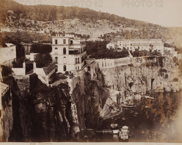 Sorrento, View of the cliffs of Sorrento, Italy, photograph, number: '2129', photographer: Giorgio Sommer, Naples, 1857 - 1914, paper, albumen print, albumendruk, height 201 mm x width 253 mm, city view in general, 'veduta', coastal cliff or crag, A wide sepia-toned view shows a large multi-story building with balconies and arched windows perched atop a rocky cliff, surrounded by terraces, stone walls, and vegetation; adjacent structures and courtyards extend along the cliff edge toward a cluster of lighter-colored buildings set back among trees; steep, jagged rock faces descend to a narrow shoreline with scattered boulders and a small wooden pier supporting a modest boathouse; paths and stairways are visible cut into the cliff and connecting levels, while the distant hillside rises with dense tree cover and scattered small buildings.