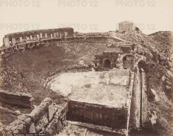 Remains of the amphitheater of Taormina, Italy, Interno del Teatro, Taormina, photograph, number: 'No. 1744.', 1851 - 1900, photographer, Sicily, pasteboard, paper, albumen print, height 309 mm x width 385 mm, ruin of a building, architecture, amphitheatre, A sepia-toned aerial view of a ruined stone structure on a rocky hillside showing curved and straight masonry walls, terraces, and arched openings, with overgrown vegetation filling some of the excavated areas; a small standalone rectangular building sits near the top of the ridge with a path leading to it, and scattered stones and rubble lie across the scene.