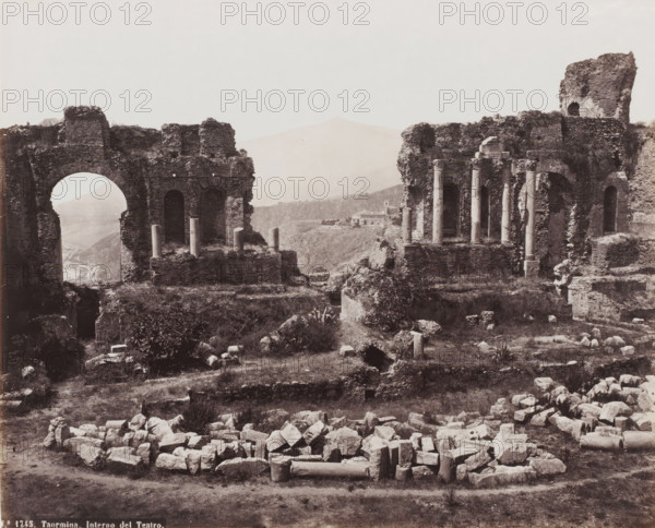 Interno del Teatro, Remains of the Amphitheater of Taormina, Italy, Taormina, photograph, number: 'No. 1745.', 1851 - 1900, photographer, Sicily, pasteboard, paper, albumen print, height 310 mm x width 384 mm, ruin of a building, architecture, amphitheatre, A wide sepia photograph shows ancient stone ruins with large arched openings and sections of crumbled walls, some topped with short columns and scattered column drums lying on the ground; the foreground contains a pile of irregularly shaped stones and broken architectural fragments on grassy earth, while midground terraces and raised platforms support remnants of masonry and upright columns; distant hills and a faint mountain silhouette appear through an archway and between the ruins, and a small cluster of buildings sits on a distant ridge; vegetation including low bushes and patches of grass grows among the ruins, and the sky above is pale and clear.