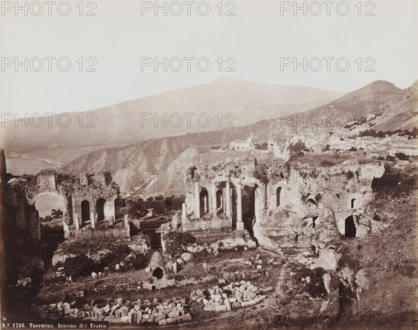 Remains of the amphitheater of Taormina, Italy, Interno del Teatro, Taormina, photograph, number: 'No. 1746.', 1851 - 1900, photographer, Sicily, pasteboard, paper, albumen print, height 310 mm x width 385 mm, ruin of a building, architecture, amphitheatre, A wide sepia-toned landscape showing ancient stone ruins with tall arches and freestanding columns in the foreground, scattered stone blocks and broken column segments on uneven ground, terraced rocky terrain with patches of vegetation, deeper ruins with arched openings and crumbling walls, a distant hillside village of light-colored buildings clustered along slopes, a small churchlike building with a simple tower among the village structures, rolling hills and a large mountain rising in the background under a pale sky.