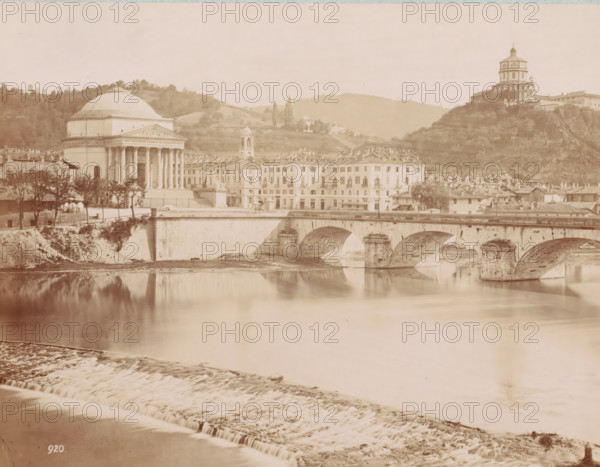 Bridge over the Po at Turin, Italy, Ponte sul Po, Torino, photograph, inscription: 'Turin, the bridge over the Po.', number: '920.', Turin, 1851 - 1900, photographer, Piedmont, pasteboard, paper, albumen print, height 257 mm x width 338 mm, bridge in city across river, canal, Po, A riverscape with a stone bridge featuring multiple arches spanning calm reflective water, a stepped weir in the foreground, a domed neoclassical building with columns near the riverbank, a row of multi-story buildings with uniform facades and tiled roofs beside it, a bell tower rising behind the row, trees and vegetation along the bank and hills in the background, and a hilltop building with a layered tower overlooking the scene.