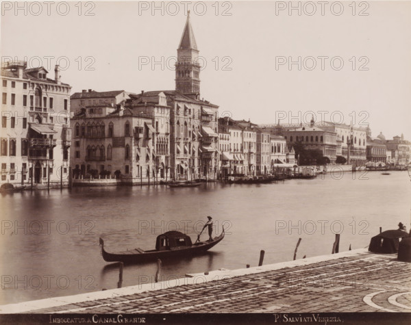 Imboccatura, Grand Canal, View of the entrance to the Grand Canal in Venice, photograph, inscription: 'Venetie', photographer: Paolo Salviati, Venice, 1860 - 1870, pasteboard, paper, albumen print, albumendruk, height 196 mm x width 250 mm, height 275 mm x width 364 mm, canals, waters (in city), gondola, A waterfront scene showing a row of multi-story buildings with varied facades, balconies, awnings, and arched windows lining a canal, with a tall bell tower rising behind them; calm water occupies the foreground where a gondola with a standing gondolier and a covered cabin floats, another gondola prow and a person are partially visible at the lower right, and a paved quay with patterned stone and wooden mooring posts runs along the bottom edge.