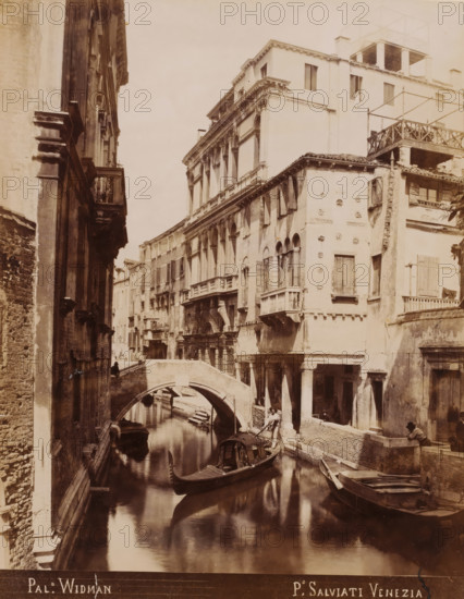 View of the Palazzo Rezzonico-Widmann in Venice, Pal. Widman, photograph, inscription: 'Venetie', photographer: Paolo Salviati, Venice, 1860 - 1870, pasteboard, paper, albumen print, height 250 mm x width 197 mm, height 348 mm x width 275 mm, urban housing, canals, waters (in city), bridge in city across river, canal, A narrow canal flanked by tall, weathered buildings with arched windows and small balconies runs under a stone bridge, with a covered gondola and other small boats on the calm water and a gondolier standing in one boat, while people sit or stand along the canal edge near columned porticos and shuttered windows, and the facades show ornamental stonework, worn plaster, and rooftop terraces.