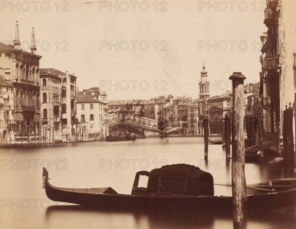 View of the Grand Canal with the Rialto Bridge in the background in Venice, Italy, photograph, Venice, 1851 - 1900, photographer, pasteboard, paper, albumen print, height 187 mm x width 240 mm, urban housing, canals, waters (in city), bridge in city across river, canal, Grand Canal, Rialto Bridge, A sepia-toned view of a canal framed by rows of multi-story buildings with arched windows and balconies on both sides, a stone bridge spanning the canal in the middle distance, a domed bell tower rising beyond the bridge, wooden posts and mooring poles along the water's edge, and a gondola with an ornate prow and covered passenger cabin in the foreground.
