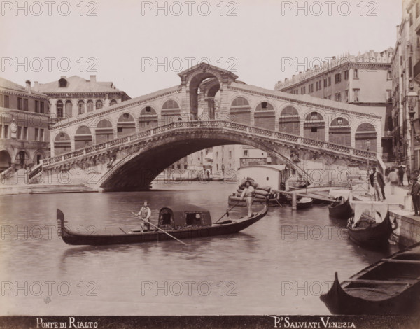 Gondolas on the Grand Canal in front of the Rialto Bridge in Venice, Ponte di Rialto, photograph, inscription: 'Venetie', photographer: Paolo Salviati, Venice, 1860 - 1870, pasteboard, paper, albumen print, albumendruk, height 198 mm x width 251 mm, height 275 mm x width 364 mm, bridge in city across river, canal, gondola, Rialto Bridge, A wide stone arch bridge with decorative balustrades spans a canal, flanked by multi-story buildings with arched windows and shuttered openings; gondolas and small boats float on the water beneath the bridge, one with a standing oarsman and another with a seated oarsman, and a small dock area with piled sacks and a person near the water is visible to the right while several figures stand or walk along the waterfront steps and quays.