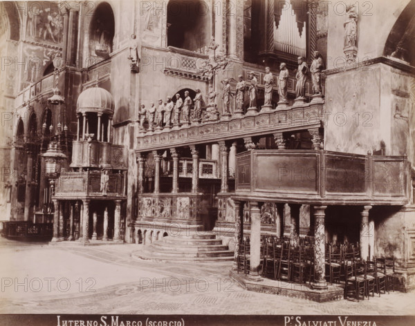 Interior of the San Marco in Venice, Interno S. Marco (scorgio), photograph, inscription: 'Venetie', photographer: Paolo Salviati, Venice, 1860 - 1870, pasteboard, paper, albumen print, albumendruk, height 197 mm x width 250 mm, height 275 mm x width 365 mm, parts of church interior, St. Mark's Basilica, A stone interior with ornate carved railings and a raised platform accessed by semicircular steps, supported by columns with decorative capitals and marbleized shafts, a row of statues standing on pedestals along the balustrade, an elevated domed pulpit with slender columns and a sculpted figure beneath it, hanging lamps and a large freestanding decorative lampstand, multiple wall niches and arches, an organ case with visible vertical pipes recessed into the wall, carved reliefs and patterned panels on the lower fronts, clustered wooden chairs lined beneath the gallery, mosaic flooring with geometric patterns, and tall wall-mounted statues and sculptural details on the surrounding surf...