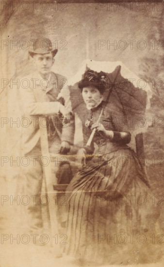 Portrait of an unknown woman with parasol and an unknown man, photograph, carte-de-visite, 1851 - 1900, photographer, inorganic material, glass, paper, albumen print, with glass cover plate, height 104 mm x width 62 mm, height 109 mm x width 69 mm x depth 4 mm, adult woman, adult man, anonymous historical persons depicted in a double portrait, parasol, sunshade, A sepia photograph of a man and a woman posed together; the man stands to one side wearing a jacket and a hat with one arm resting on a wooden prop, while the woman sits holding a parasol above her shoulder and wears a dark dress with a high collar, gloves, and a decorated hat. The woman's skirt has visible vertical pleats and texture, and the parasol has a scalloped edge. Both faces are visible with neutral expressions. The background is plain with a mottled studio backdrop, and the image shows signs of age such as fading and small spots.