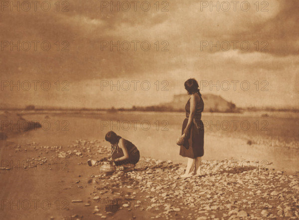 On the Rio Grande- San Ildefonso, photograph, photographer: Edward Sheriff Curtis, 1900, photoaquatint, heliogravure, height 450 mm x width 560 mm, height 293 mm x width 392 mm, A sepia-toned scene of a shoreline with a cloudy sky and distant land across calm water, showing pebbles and small rocks along the wet shore; two women are present, one standing with short dark hair wearing a sleeveless dark dress and holding a rounded object at her side while looking toward the water, the other crouching near the water's edge with dark hair tied back and wearing a dark top and lighter skirt while handling a large vessel placed on the ground.