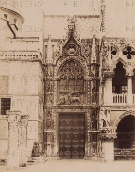 Porta della Carta in the Basilica of San Marco in Venice, Italy, Porta della Carta, Venezia, photograph, number: '5.', Venice, 1851 - 1900, photographer, pasteboard, paper, albumen print, albumendruk, height 369 mm x width 322 mm, parts of church exterior and annexes: portal, Florence, Tuscany, San Marco, A sepia-toned photograph of an ornate stone building facade showing a large wooden door with raised rectangular panels set within a richly carved archway decorated with tracery and circular floral motifs; flanking the doorway are sculpted columns and niches containing standing human figures, and above the arch is a relief of a winged lion next to a robed kneeling figure beneath a circular medallion containing a sculpted bust, all set among elaborate foliage and architectural ornament; to the right a section of arcade with rounded arches and quatrefoil openings is visible, with additional sculpted figures and ornate capitals, while to the left plain stone walls and a small barred windo...