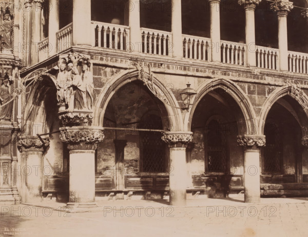 Angolo di P. Ducale, Detail of the Doge's Palace in Venice, Italy, Venezia, photograph, number: '24.', Venice, 1851 - 1900, photographer, pasteboard, paper, albumen print, albumendruk, height 321 mm x width 370 mm, architectural detail, gallery (spacious corridor), Doge's Palace, A sepia photograph showing an ornate stone arcade with tall columns and pointed arches, carved capitals with foliage motifs, and sculpted figures on a corner capital; a decorative balustrade runs above the arches between columns, with a dark recessed upper loggia behind it; an iron lantern hangs from one arch and scrollwork metal supports are attached to the stone; beneath the arches are dark openings and windows with diamond-patterned grilles, and wooden benches or ledges along the interior wall; the stone pavement in front is sunlit with soft shadows cast by the columns.