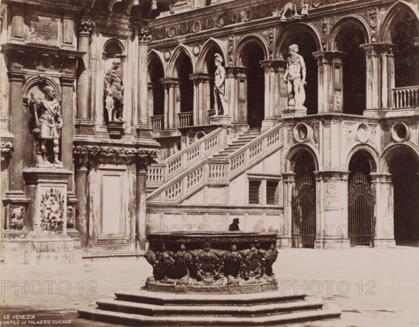 Courtyard of the Doge's Palace in Venice, Italy, Cortile del palazzo Ducale, Venezia, photograph, number: '68', Venice, 1851 - 1900, photographer, pasteboard, paper, albumen print, albumendruk, height 319 mm x width 368 mm, inner court of palace or 'hôtel', Doge's Palace, A stone courtyard scene showing a large ornate circular fountain basin on a stepped platform in the foreground, the basin decorated with sculpted faces and ornamental reliefs. Behind it a grand exterior wall of a palatial building with carved columns, arched openings, decorative medallions, and balustraded stairs leading up to a terrace. Several life-sized stone statues stand in niches and on pedestals along the facade, including figures in classical poses and draped garments, and one bearded figure. There are wrought iron gates set into arched doorways and carved panels and reliefs filling many of the architectural surfaces. A solitary figure sits on the edge of a raised platform near the base of the staircase. The i...