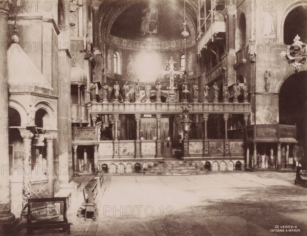 Interno S. Marco, Interior of San Marco in Venice, Italy, Venezia, photograph, number: '131', Venice, 1851 - 1900, photographer, pasteboard, paper, albumen print, albumendruk, height 320 mm x width 365 mm, interior of church, Florence, Tuscany, San Marco, A sepia-toned interior view of a large ornate space dominated by a raised altar area with a balustrade topped by a row of standing statues and a central decorative cross, framed by columns and arches; behind the altar a semicircular apse with painted figures and small arched windows rises beneath an embellished arch, while scaffolding and a suspended lamp are visible to one side; the foreground shows a wide open floor with worn stone paving, early wooden benches along the left, carved pillars and arched openings, and various carved reliefs and architectural details on the walls and cornices throughout the scene.