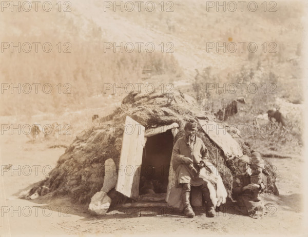 Family in front of their home (gamme) in Sápmi, Family in front of a round hut in Lapland (former title), photograph, Sápmi, 1860 - 1900, photographer, paper, albumen print, height 162 mm x width 208 mm, family life, Europeans: Sami, A small earthen shelter with a rounded, grass-covered roof and a wooden plank propped at its entrance is set against a pale, textured landscape; a man in layered outer garments and a headwrap sits on the shelter's stone threshold holding a light-colored animal pelt, while two children in patterned clothing and head coverings sit on the ground beside him, one child appearing to hold something in their lap; rocks and scattered vegetation surround the shelter and a grazing animal is visible farther back in the scene.