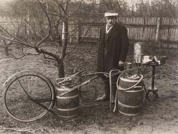 Man by a self-built pesticide spraying system for fruit trees, possibly in Denmark, photograph, Denmark, 1900 - 1930, photographer, paper, baryta paper, gelatin silver print, developing gelatin silver print, height 171 mm x width 228 mm, adult man, fruit-tree culture, fruit harvest, A person wearing a long dark overcoat, layered clothing, boots, and a hat stands near wooden barrels and a small round table holding metal cans and a few tools, with hoses and a hand-operated pump attached to the barrels; a bare-branched tree and a wooden fence form the background, and a bicycle wheel and frame lie on the ground among scattered leaves and dirt.
