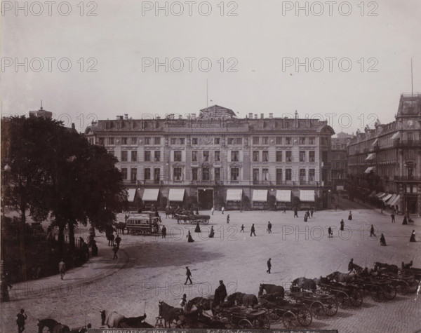 Hotel d'Angleterre, View of Hotel d'Angleterre at Kongens Nytorv in Copenhagen, Kjøbenhavn og Omegn, photograph, number: 'Nr. 43.', Copenhagen (city), publisher: Vilhelm Trydes, Denmark, 1880 - 1910, photographer, paper, pasteboard, baryta paper, daylight collodion silver print, height 225 mm x width 283 mm, height 350 mm x width 455 mm, square, circus, hotel, hostelry, inn, Kongens Nytorv, A wide plaza scene shows a large multi-story stone building with evenly spaced windows, ornate facade details, and striped awnings over ground-level openings, flanked on one side by a row of connected buildings and on the other by tall trees. Horses hitched to covered carriages are lined up along the foreground with drivers nearby. Several groups of pedestrians in period clothing are scattered across the cobbled square, some walking and some standing, while a horse-drawn omnibus is visible near the left side of the plaza. The sky above is open and pale, and architectural roof details and statues lin...
