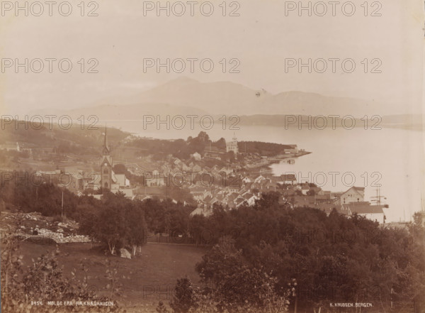 View of Molde in Norway, Molde from Ræknæshaugen, photograph, number: '8454.', photographer: Knud Knudsen, Molde, 1888, paper, albumen print, albumendruk, height 161 mm x width 216 mm, city-view in general, 'veduta', A sepia-toned panoramic view shows a coastal town with clustered houses and larger buildings along a shoreline, a tall church with a pointed steeple rising prominently among the rooftops, tree-covered areas in the foreground and scattered trees throughout the town, a pier extending into calm water, distant low hills or mountains across the water, and an open sky above.