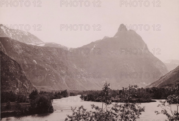 View of the Romsdalshornet in Norway, Romsdalshorn og vængetinderne, photomechanical print, number: 'No 219.', copyright notice: 'ENERET 1891.', Norway, publisher: F. Beyer, Bergen, cliché maker: anonymous, 1891, photographer, paper, collotype, photoprint, height 154 mm x width 212 mm, mountains, Romsdalshornet, A wide landscape showing a river flowing toward the foreground with low trees and shrubs along its banks, a broad flat plain beyond the near water, steep rocky mountain slopes rising on both sides, a prominent jagged peak on the right with patches of snow on its upper ridges, additional serrated snowy ridges visible on the left, and a pale sky above.