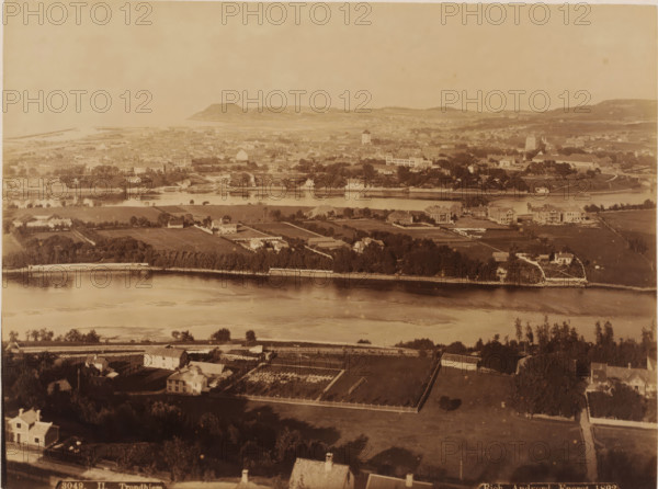Trondhiem, View of Trondheim and the Nidelva, photograph, number: '3049.', inscription: 'Eneret', Trondheim, publisher: Andvord, Rich., Oslo, 1892, photographer, pasteboard, paper, albumen print, albumendruk, height 168 mm x width 224 mm, height 348 mm x width 455 mm, city-view in general, 'veduta', river, Nidelva, A wide aerial view of a riverside town with a broad body of water running horizontally through the middle of the image, grassy and tree-lined banks along the near and far shores, and clusters of buildings and houses spread across fields and plots on both sides of the river; the foreground shows fenced yards, individual houses, garden plots, and tree shadows, the middle ground includes larger houses, villas, and a tree-lined strip along the far riverbank, and the background reveals a dense collection of rooftops, church-like structures, a distant shoreline with low hills, and a hazy sky above.
