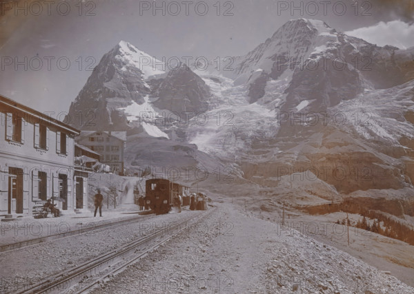 Wengernalp with Eiger and Mönch, View of a railway track in Wengen at the foot of the Eiger and the Mönch in the Bernese Alps, Hotel Jungfrau on the left, photograph, number: '3664.', Wengen, 1890 - 1930, photographer, paper, baryta paper, daylight gelatin silver print, height 164 mm x width 220 mm, railway, train, mountains, A mountainous landscape fills the scene with snow-covered peaks and exposed rock faces rising in the background, and a large glacier flowing down between steep cliffs. In the foreground a railway track runs toward the mountains with a small train stopped on the line and a group of people gathered near the cars. To the left, a multi-story building with shuttered windows lines the platform where a few individuals sit on a bench and another stands nearby; another building sits slightly back on a slope. The ground near the tracks is gravelly and uneven, and the right side of the image shows sloping terrain with scattered rocks and sparse vegetation.