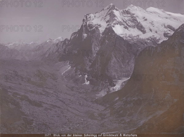 View of the Kleinen Scheidegg auf Grindelwald & Wetterhorn, View of the Grindelwald and the Wetterhorn from the Kleine Scheidegg, photograph, number: '3577.', Kleine Scheidegg, 1890 - 1930, photographer, paper, baryta paper, daylight gelatin silver print, height 164 mm x width 219 mm, mountains, Grindelwald, Wetterhorn, A wide mountainous landscape with a large snow-capped peak dominating the right side and rugged dark rock faces beneath the snow, tapering down into steep cliffs and a deep valley; patches of snow and ice sit on ledges and in gullies, and lower slopes show grassy and rocky terrain with scattered trees and small clusters of buildings visible near the valley floor; distant mountain ridges and peaks extend across the left and center background under a pale, cloudless sky.