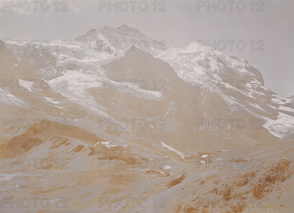 Kleine Scheidegg, View of the Kleine Scheidegg in the Bernese Alps, photograph, number: '3678.', Berner Alpen, 1880 - 1910, photographer, Alps, Bernese Alps, paper, baryta paper, daylight collodion silver print, height 165 mm x width 219 mm, mountains, A wide mountain scene showing a large rocky peak with snow and ice on its upper slopes and ridges under a pale sky, with steep cliffs and layered rock faces below; grassy and rocky foothills lead down from the mountain, where several multi-story chalet-style buildings and smaller wooden structures sit scattered along a winding road and plateau near the base of the slopes.