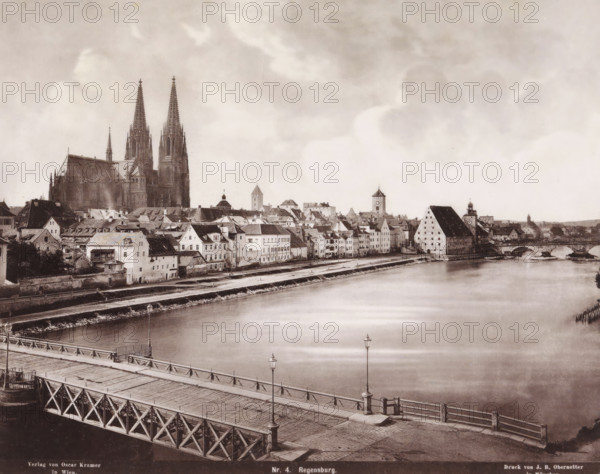 View of Regensburg, Germany, Regensburg, photomechanical print, number: 'Nr. 4.', cliché maker: Johann Baptist Obernetter, Munich, publisher: Oscar Krämer, Vienna, 1856 - 1887, photographer, Bavaria, Lower Austria, pasteboard, paper, collotype, photoprint, height 305 mm x width 372 mm, city-view in general, 'veduta', A riverside cityscape with a wide calm river in the foreground, a wooden bridge crossing the river with visible railings and lampposts, and rows of multi-story buildings with pitched roofs lining the riverbank; a large Gothic cathedral with two tall spires rises prominently behind the buildings, and several towers and a distinctive sloped-roof warehouse or granary appear among the rooftops; a stone arched bridge connects the far bank, and the sky above is filled with pale, soft clouds.