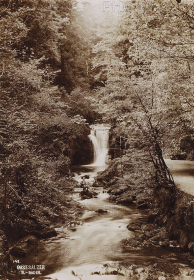 Waterfall with standing boy, photograph, date: '1903', inscription: '148., Gust. Salzer, B.-Baden.', photographer: Gustav Salzer, Baden-Baden, 1903, Baden-Württemberg, paper, baryta paper, daylight collodion silver print, height 168 mm x width 117 mm, waterfall, A sepia-toned scene of a forested ravine with a waterfall cascading into a stream that flows toward the foreground, surrounded by dense trees and undergrowth on both banks, rocky outcrops and boulders lining the watercourse, a lone person wearing a hat seated on a rock near the stream below the waterfall, and a narrow path visible along the right bank beside leaning and twisted tree trunks.