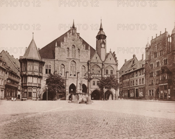 Hildesheim Town Hall, photomechanical print, inscription: 'Hildesheim', number: 'N.1.', photographer: E. Mertens & Cie (possibly), Hildesheim, cliché maker: anonymous, 1880 - 1900, Lower Saxony, paper, collotype, photoprint, height 228 mm x width 286 mm, square, circus, townhall, A wide cobblestone square fills the foreground leading to a row of ornate stone and timber buildings along the back edge. Prominent in the center is a large Gothic stone building with pointed arched openings at ground level, tall lancet windows above, stepped gables, and a clock tower with a spire rising to the right. In front of this building stands a sculpted fountain with a central column and a decorative lamp post nearby. To the left a multi-story bay with a conical roof and patterned window panes projects over the street, flanked by smaller timber-framed buildings. To the right of the central structure are steeply pitched tiled roofs and a tall stone building with narrow arched windows and decorative roof...