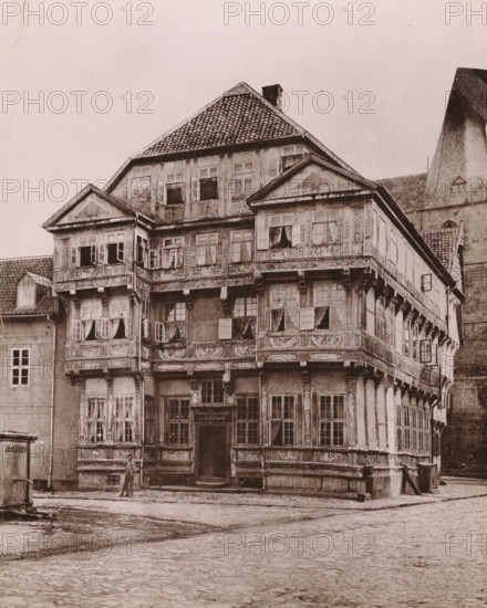 Cafe and lodging in Hildesheim (former title), Brewery and drinks room of G. Ehrhardt, Hildesheim, photomechanical print, inscription: 'N.13', inscription: 'Hildesheim', photographer: E. Mertens & Cie (possibly), cliché maker: anonymous, 1880 - 1900, Lower Saxony, paper, collotype, photoprint, height 275 mm x width 219 mm, urban housing, brewer, A multi-story timber-framed building with decorative carved panels and overhanging upper floors sits at a cobblestone street corner, its steep tiled roof and chimney visible above a facade of many small-paned windows, several of which are open or have curtains; ornate brackets support the projecting sections and a central recessed doorway is flanked by tall windows, while adjacent buildings and a tall stone structure with a steep roof rise nearby and a single person stands on the pavement in front of the building.