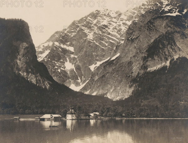 St. Bartholomew's Church on the western shore of the Königssee near Berchtesgaden, in the background the Watzmann, photograph, photographer: H. Gurtler, Berchtesgaden, 1927-08, H. Gurtler, photographer, Bavaria, paper, baryta paper, gelatin silver print, developing gelatin silver print, height 200 mm x width 260 mm, lake, church (exterior), mountains, Watzmann, Königssee, St. Bartholomew's Church (Berchtesgaden), A wide mountain scene with steep rocky peaks and patches of snow rising behind a dense dark forest, a calm reflective lake in the foreground, a small cluster of light-colored buildings with pitched roofs and a domed chapel at the water edge, a lone small boat with a person rowing on the lake, and the mountains and buildings reflected on the lake surface.