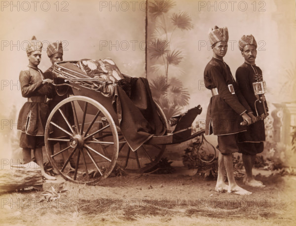 Studio portrait of four Indian men in uniform with a carriage, India, photograph, inscription: '£1', inscription: 'Asia', signature: 'SH Dagg', photographer: Sidney Herbert Dagg, British India, 1890 - 1910, pasteboard, paper, albumen print, height 213 mm x width 275 mm, height 227 mm x width 293 mm, clothes for official occasions, rickshaw, A sepia-toned photograph showing a large wooden two-wheeled carriage draped with patterned fabric and a dark cloth, flanked by four men in matching dark uniforms and tall patterned turbans; two men stand near the rear wheel holding the carriage frame while two stand at the front holding the shafts, one of them carrying a box-like lantern attached to the shaft, and all four are barefoot on a grassy surface with a painted backdrop of tall, leafy plants behind them.