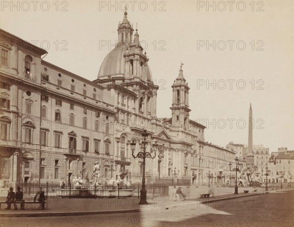 Piazza Navona, View of the Piazza Navona in Rome, Italy, Roma, photograph, number: '12-', number: '8', number: '3633.', photographer: Giacomo Brogi (possibly), Rome, publisher: attributed to Edizioni Brogi, 1864 - 1881, Attributed to, pasteboard, paper, albumen print, albumendruk, height 276 mm x width 375 mm, square, circus, A wide sepia photograph of a city square dominated by a large ornate church facade with a prominent central dome and flanking towers, showing carved stone details, columns, and arched windows; in front of the church is an ornate fenced fountain with sculpted figures and water basins, and decorative street lamps with multiple lanterns line the paved square; to the left a long row of multi-story buildings with shuttered windows and balconies stretches along the street; a tall obelisk rises in the distance to the right against a pale sky; several blurred figures and seated people appear on benches near the foreground, and shadows fall across the cobblestone pavement.