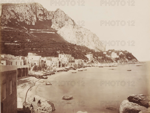 View of Marina Piccola in Capri, Italy, Capri Marina, photograph, number: '1173', inscription: 'Capri Marina', photographer: Giorgio Sommer, Capri, Isola di, 1857 - 1914, pasteboard, paper, albumen print, height 308 mm x width 385 mm, gulf, bay, A wide coastal scene showing a row of low, light-colored buildings with arched openings and flat roofs lining a narrow beach where numerous small wooden boats rest on the sand and at the water’s edge, a calm expanse of water stretches outward with several small boats floating, rocky outcrops and larger boulders appear in the foreground near the shoreline, a steep, rugged hillside and large cliff rise behind the buildings with terraced vegetation and scattered structures on the slope, and a headland with additional buildings extends into the sea on the right under a pale, empty sky.