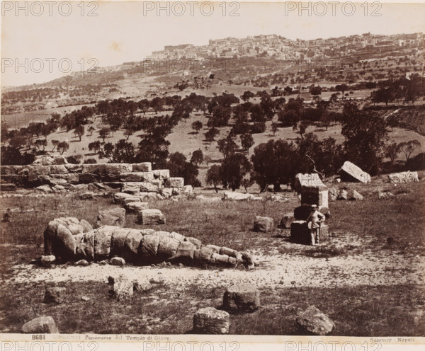 View of Agrigento from the Temple of Jupiter, Italy, Panorama dal Tempio di Giove, Girgenti, photograph, number: '8681', photographer: Giorgio Sommer, Agrigento, 1857 - 1914, Sicily, pasteboard, paper, albumen print, height 320 mm x width 369 mm, city-view in general, 'veduta', dale, valley, A sepia-toned landscape showing a hillside town in the distance above a gently sloping plain dotted with scattered trees and low vegetation, with large rough-cut stone blocks and fragments scattered across the foreground and a stepped stone structure extending across the middle ground, and a solitary person standing beside a rectangular stone block near the right side of the image.