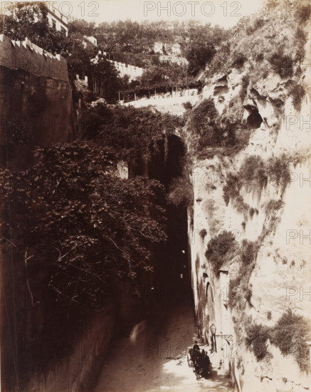 Piedigrotta, View of a bridge in Piedigrotta in Naples, Italy, Napoli, photograph, number: '1125', photographer: Giorgio Sommer, Naples, 1857 - 1914, pasteboard, paper, albumen print, albumendruk, height 384 mm x width 308 mm, rocks, bridge, A narrow sunlit pathway runs between high rock walls, the right wall showing rough pale stone with patches of grass and small caves and the left wall darker and shadowed with dense tree foliage hanging over it; near the lower right a small carriage with a driver and a passenger is parked beside the stone wall under an ornate lamp bracket, and small arched openings are visible in the right wall above them; higher up the scene shows terraced vegetation, a low white fence and a pale building partially hidden among trees on the hillside.