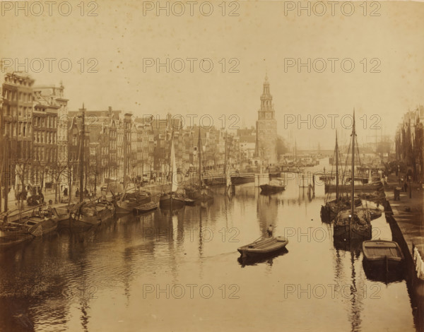 Oude Schans and Montelbaan Tower in Amsterdam, photograph, photographer: Albert Greiner, Amsterdam, 1861 - 1889, pasteboard, paper, albumen print, height 268 mm x width 342 mm, height 484 mm x width 654 mm, urban housing, canal, ships (in general), quay, Oudeschans, Montelbaan Tower, A sepia-toned city scene showing a canal lined with narrow multi-story buildings on both sides, with numerous moored sailboats and rowboats along the water edges and reflections on the canal surface, a small rowboat with a person rowing toward the foreground, a stone bridge crossing the canal further back, and a tall clock tower rising near the bridge with streets and people visible along the quay.