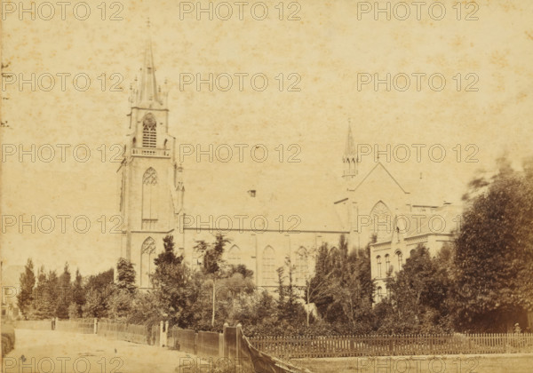 View of the Immaculate Conception of Our Lady in Overveen, View of the St. Bavo Church in Haarlem (former title), Environs de Haarlem, cabinet photograph, number: '25', publisher: Andries Jager, Amsterdam, 1860 - 1890, photographer, paper, pasteboard, albumen print, height 109 mm x width 168 mm, church (exterior), Haarlem, St. Bavo Church (Haarlem), A sepia-toned photograph showing a large Gothic-style stone church with tall pointed spires and arched stained-glass windows partially obscured by trees in the foreground, a picket fence running along a dirt road in front of the trees, dense foliage and shrubs occupying the lower half of the image, architectural details on the church including buttresses and ornamental window tracery, a smaller attached building with rectangular windows to the right, and a pale, textured sky filling the upper portion of the scene.