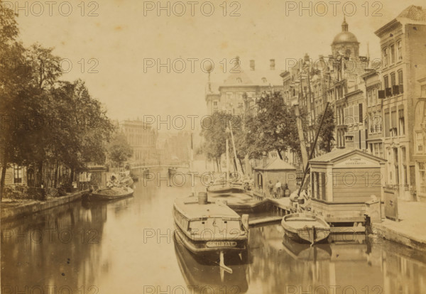 The Pipe Market, View of the Pipe Market in Amsterdam, Amsterdam, cabinet photograph, annotation: 'NZ Voorburgwal - pipe market, and old flower market, filled in 1884.', publisher: Andries Jager, 1860 - 1884, photographer, paper, pasteboard, albumen print, height 109 mm x width 167 mm, canals, waters (in city), A narrow waterway runs through the scene with several moored boats of varying sizes along the right bank and one large vessel prominent in the foreground, its bow reflected in the water; wooden docks, barrels, and small boathouses line the right edge of the canal. Tall, narrow multi-story buildings with shuttered windows and decorative facades rise behind the docks, interspersed with leafy trees whose canopies extend over parts of the canal. On the left bank a row of trees and low buildings create a shaded promenade with a small boat tied up at the edge. A bridge spans the canal in the mid distance beyond which larger stone buildings and architectural details are visible, and a ...